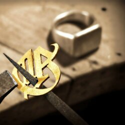 Goldenes handgesägtes Monogramm auf Laubsäge in Goldschmiede-Werkstatt, im Hintergrund unscharfer Siegelring-Rohling